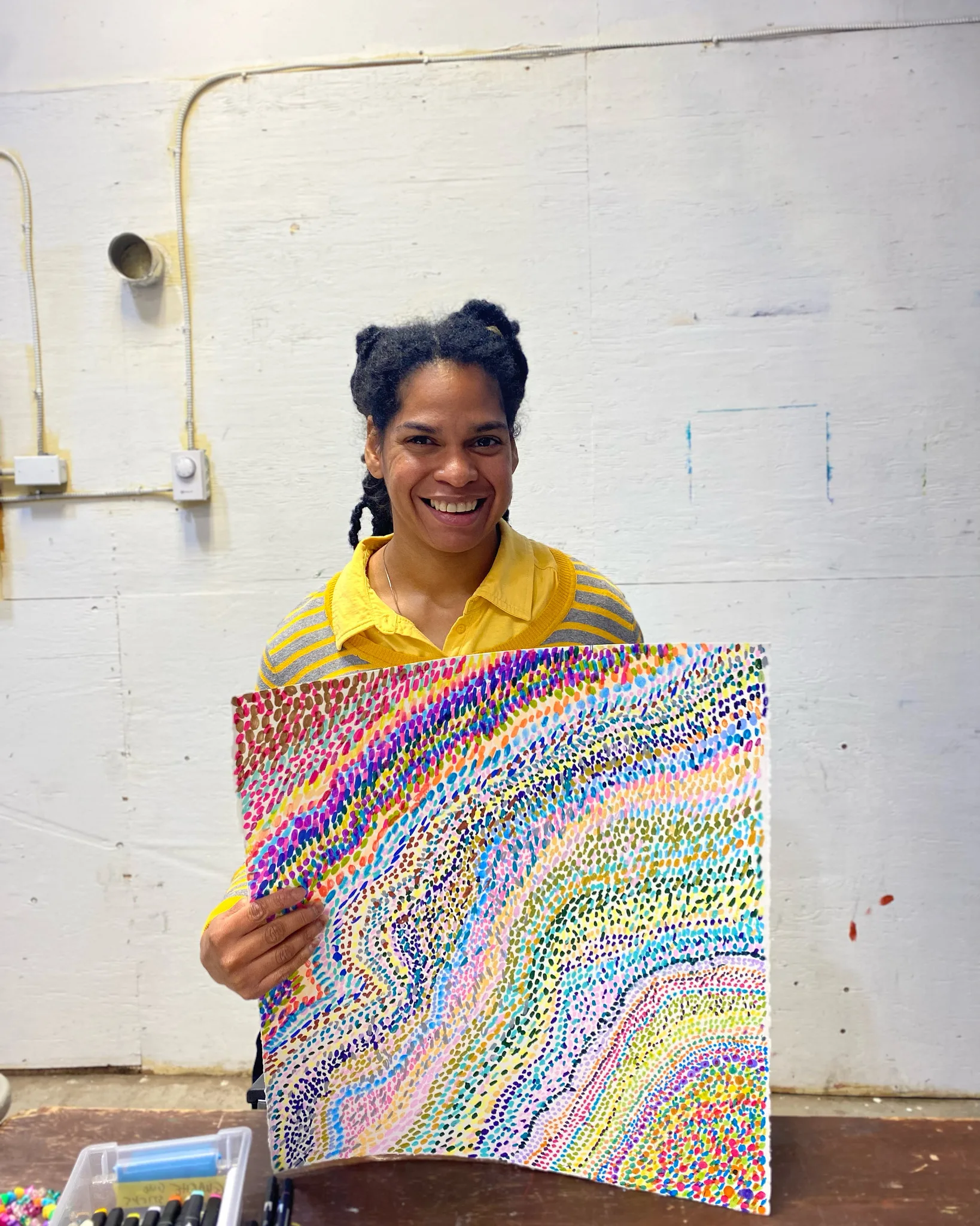 Anna smiling while holding a colourful piece of artwork made up of flowing rows of small painted dots, 2025