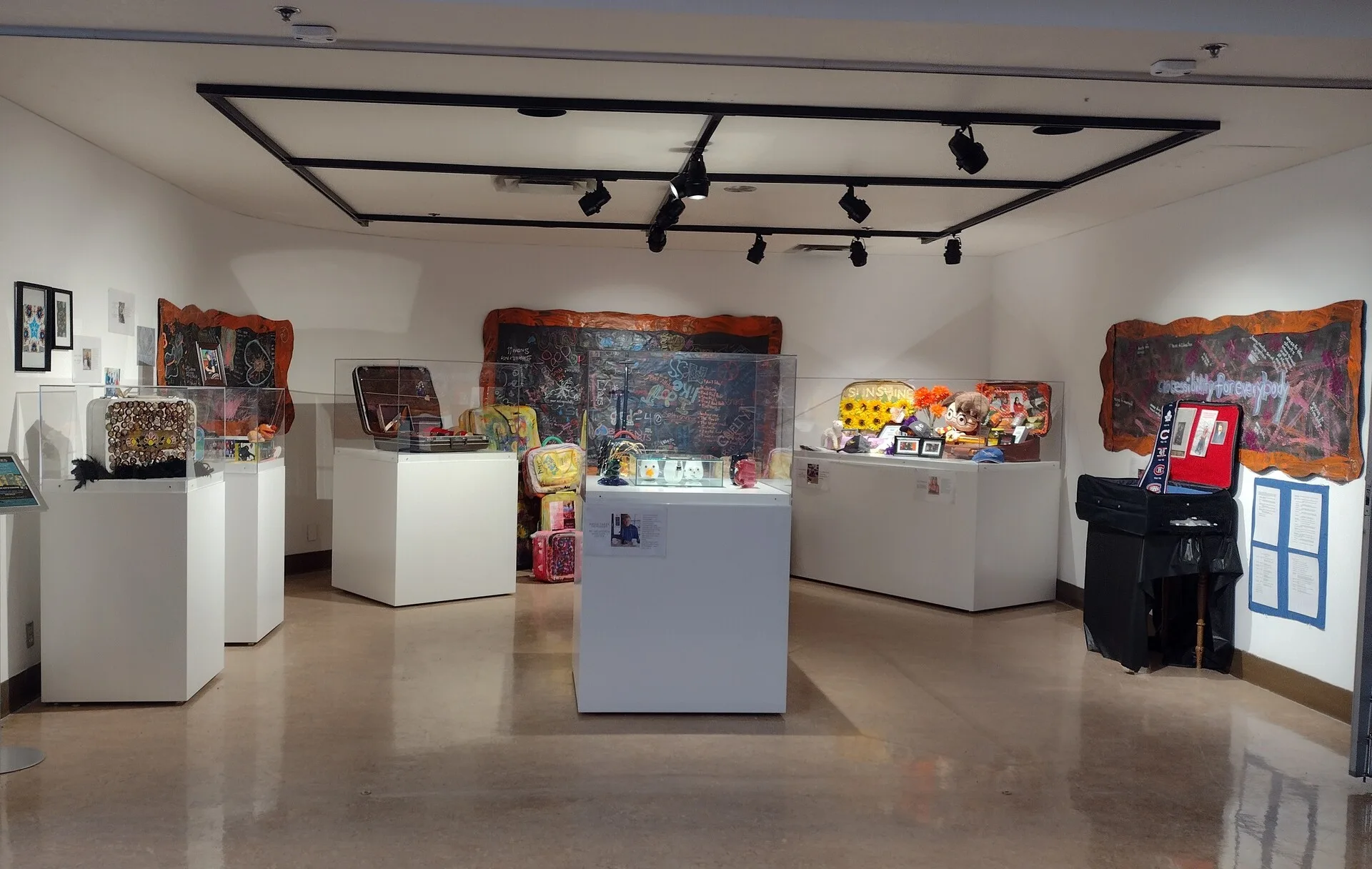 Art exhibition featuring decorated suitcases, sculptures, framed artworks, and mixed-media pieces displayed on white pedestals and gallery walls, 2022