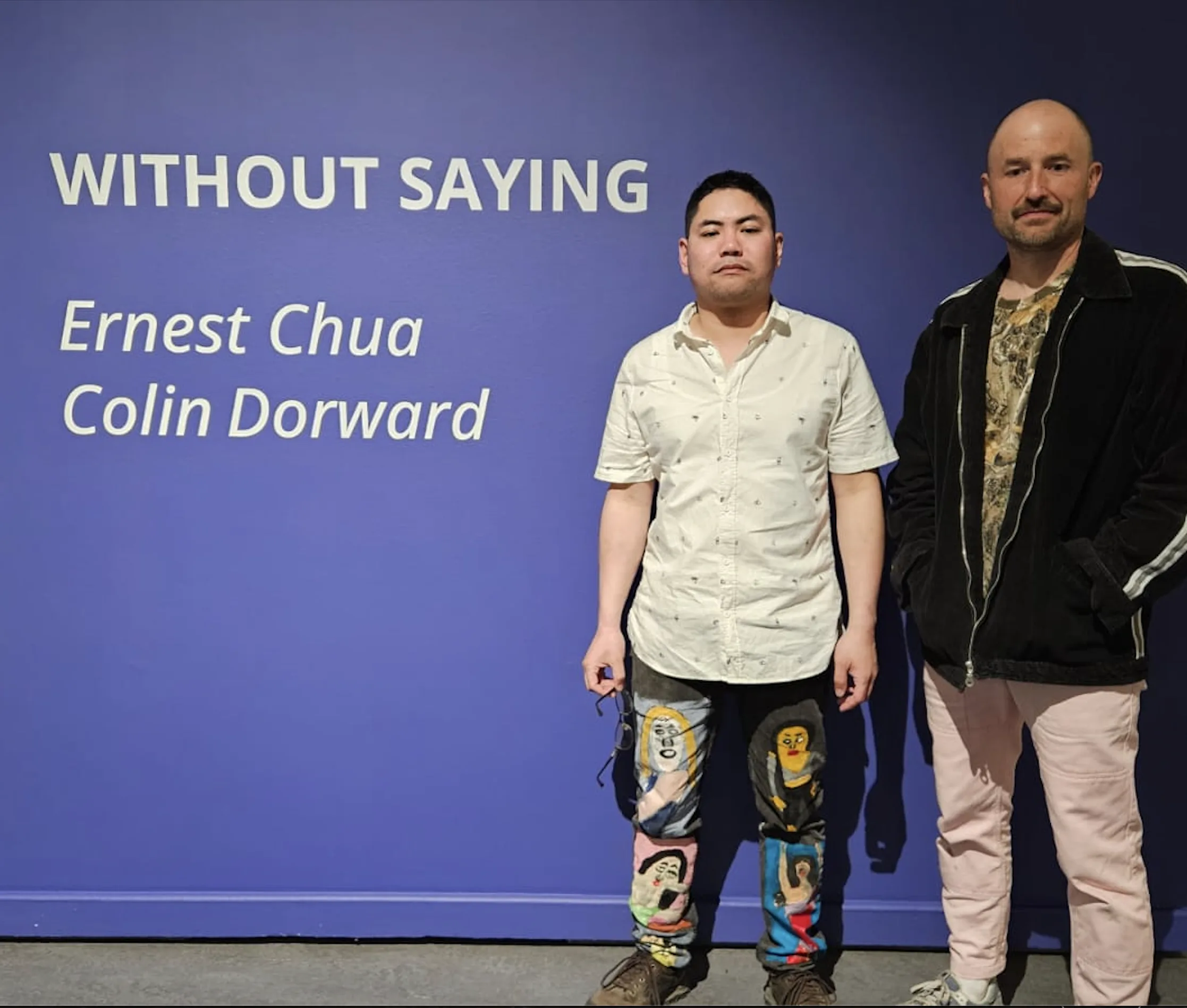Promotional image of Ernest and Colin standing in front of signage for their exhibit, Without Saying