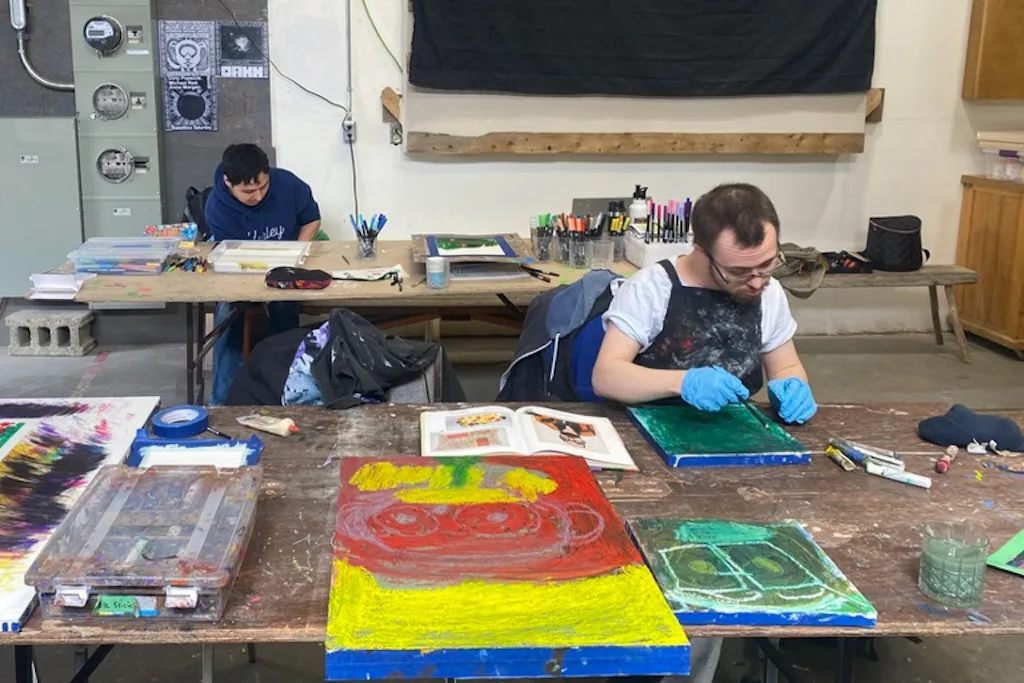 Aaron creating art at the Warehouse, with colourful artwork and supplies spread across the table, 2025
