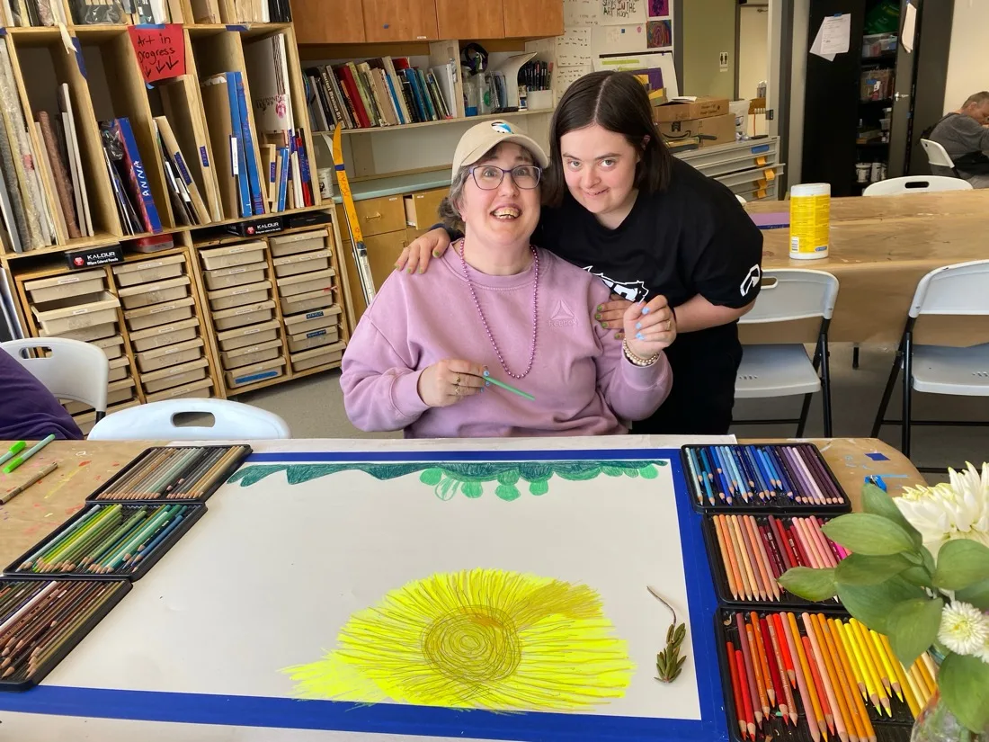 Theresa and Tijana smiling together at the Progressive Art Studio while Theresa works on a large colourful drawing with coloured pencils, 2026