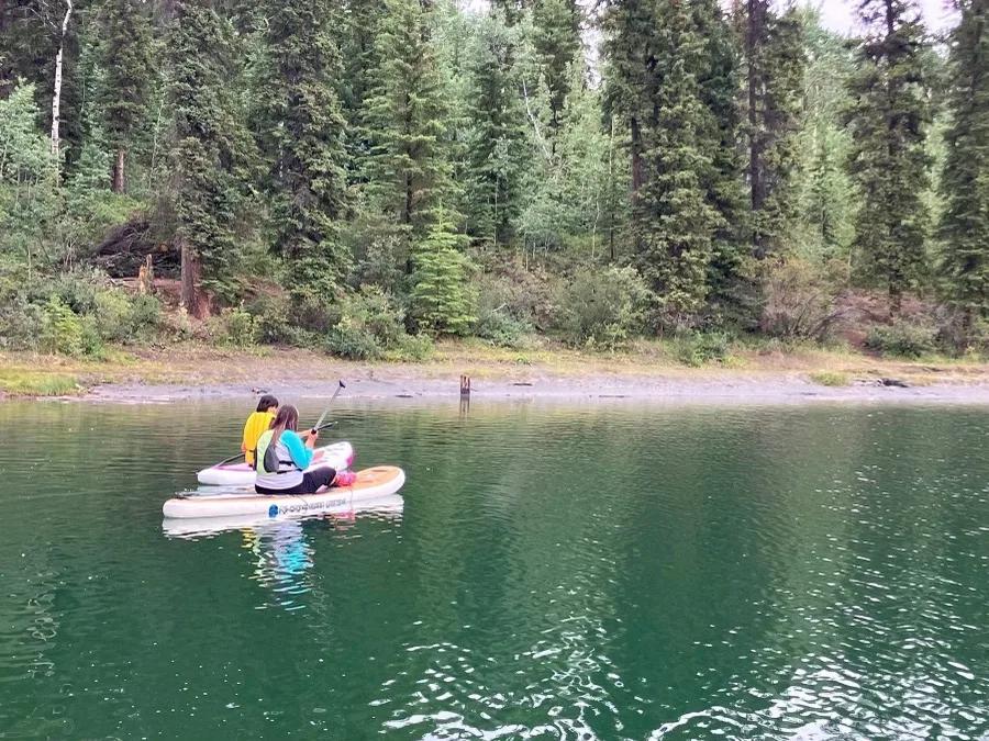 Taylor and Rachel paddleboarding together on a calm lake surrounded by dense forest, 2025
